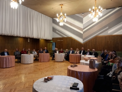 Personas sēž pie apaļiem galdiem lielā zālē