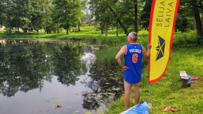 Laivotājs sporta tērpā stāv ūdens tilpnes krastā ar zāli, blakus zila kanoe laiva