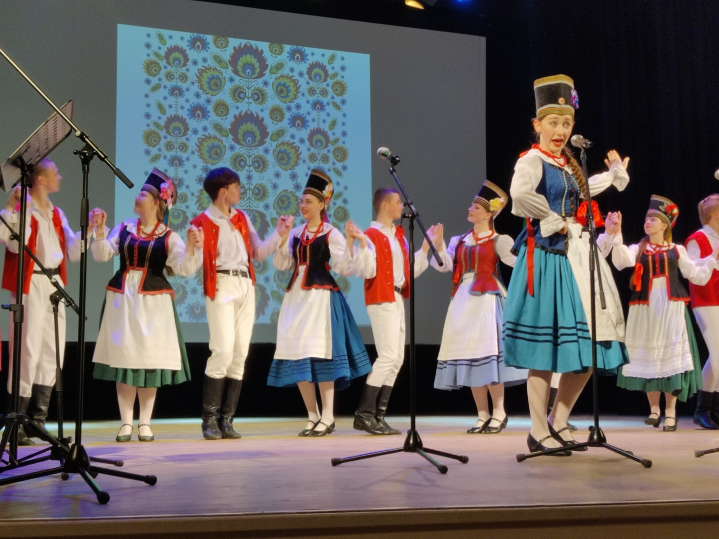 Uz skatuves stāv cilvēki poļu tautastērpos, priekšplānā sieviete spēlē instrumenti