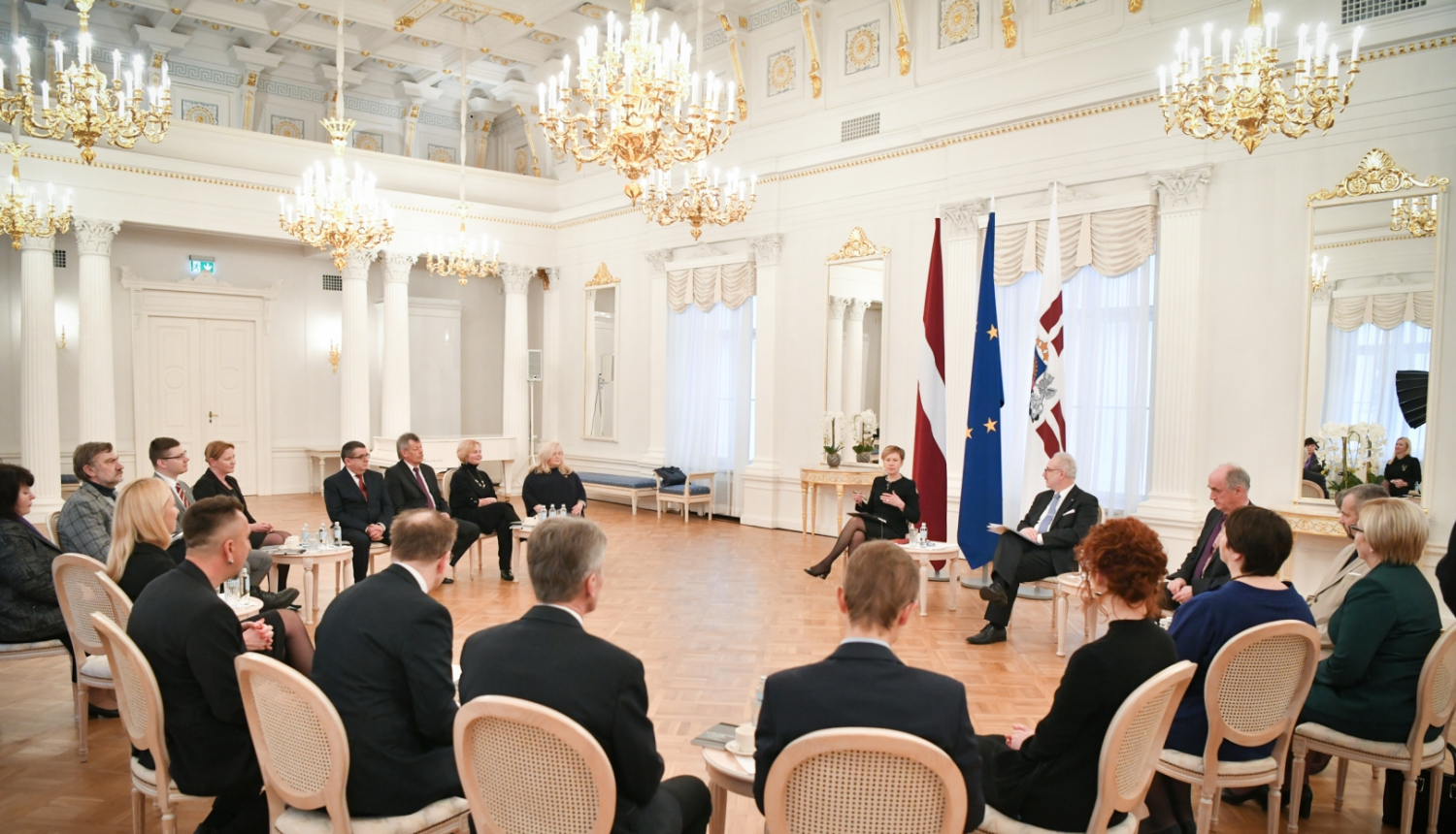Personas sēž aplī krēslos lielā greznā baltā zālē pie prezidenta