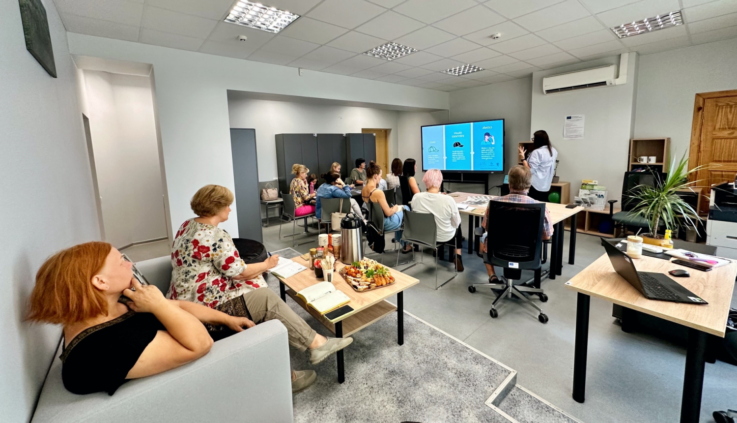 Foto no apmācībām - personas sēž pie galdiem telpā un skatās ekrānā uz lektora norādīto