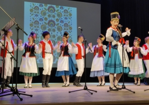 Uz skatuves stāv cilvēki poļu tautastērpos, priekšplānā sieviete spēlē instrumenti