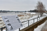 Ziemas skatā priekšplānā pelēks informatīvais stends ar tekstu dažādās valodās, fonā Daugava un Jēkabpils tilts