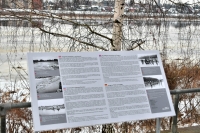 Ziemas skatā priekšplānā pelēks informatīvais stends ar tekstu dažādās valodās, fonā Daugava un bērzs