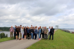 Pašvaldību dalībnieki stāv uz ceļa, fonā Daugava un HES