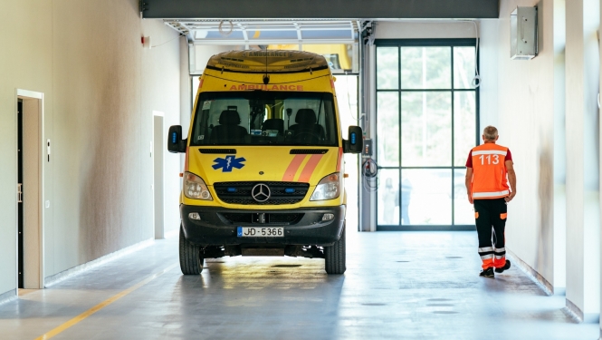 Dzeltena ātrās palīdzības mašīna telpās, blakus pa labi iet ātrās palīdzības darbinieks oranžā vestē