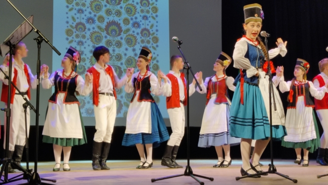 Uz skatuves stāv cilvēki poļu tautastērpos, priekšplānā sieviete spēlē instrumenti