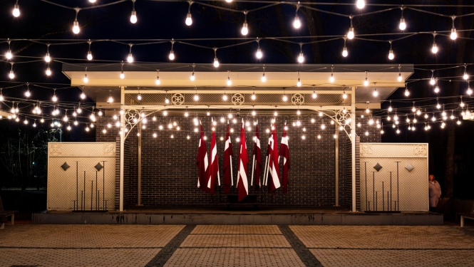 Kena parka estrāde naktī - izgaismota ar lampiņām un fonā Latvijas karogi