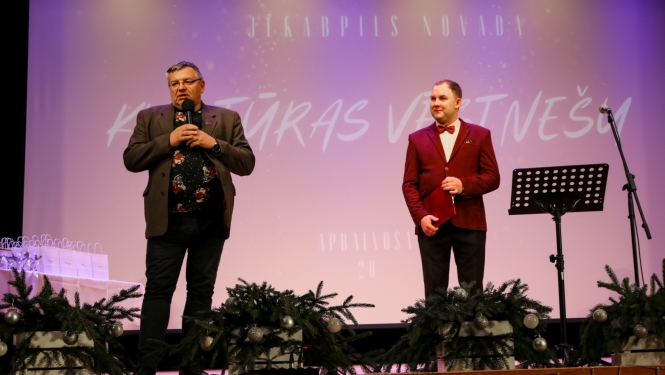 No kreisās Alfons Žuks ar vadītāju uz skatuves, fonā ekrāns ar uzrakstu Jēkabpils novada Kultūras vēstnešu apbalvošana