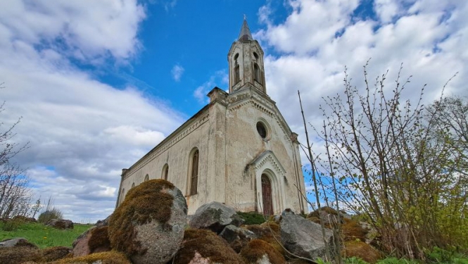 Pelēcīgi baltā Kaldabruņas baznīca pavasarī, fonā mākoņainas debesis, priekšplānā akmeņu krāvumi