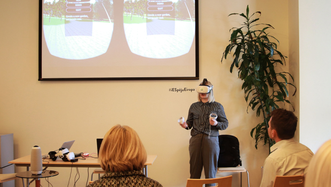 pubicitātes foto - persona telpā izmanto virtuālās realitātes brilles, fonā uz ekrāna tiek projecēts, jko persona brillēs redz