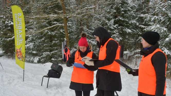 Pusstundas skrējiena ilustratīvs foto - cilvēki vestēs ziemīgā mešā skatās orientēšanās kartē