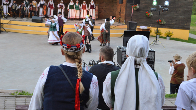 Tautumeitas no mugurpuses sēž, vēro koncertu Sēlija rotā, tālumā skatuve un dejotāji