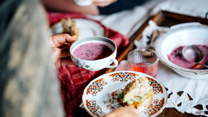Persona pie galda no mugurpuses, sēdus, rokās tur šķīvi, fonā aukstās zupas bļodiņas