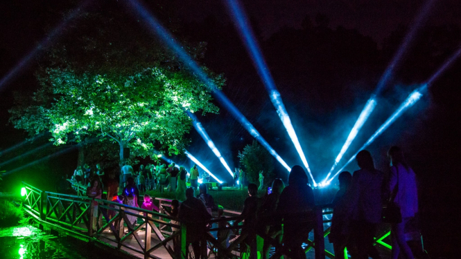 Cilvēki natī iet par tiltiņu uz saliņu, kurā debesīs spīd gaismas stari un izgaismots koks uz salas