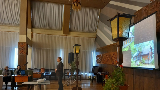 Persona runā lielā zālē, fonā projektorā rāda ēkas bildi