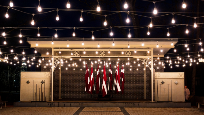 Kena parka estrāde naktī - izgaismota ar lampiņām un fonā Latvijas karogi