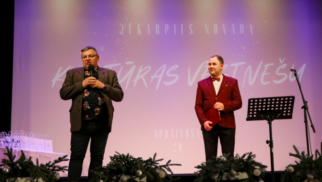 No kreisās Alfons Žuks ar vadītāju uz skatuves, fonā ekrāns ar uzrakstu Jēkabpils novada Kultūras vēstnešu apbalvošana