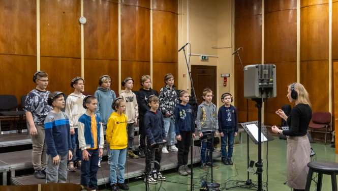 Zēnu koris sastājušies dzied un sieviete diriģē