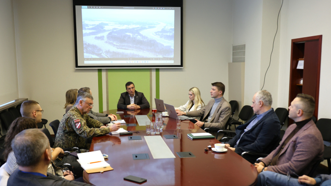 Komisija telpā pie galda, fonā ekrāns ar Daugavas skatu