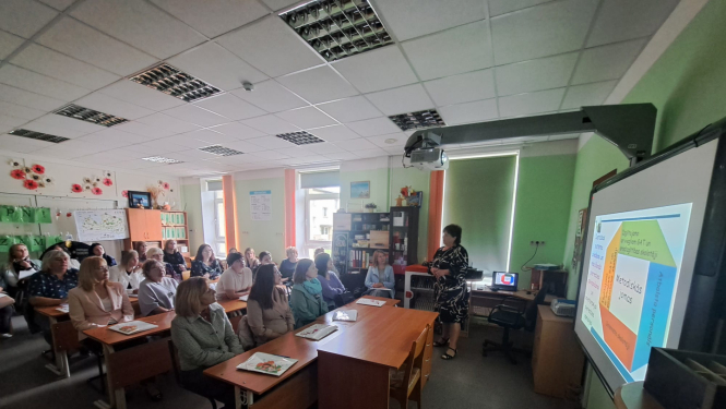 Seminārs telpās. Personas sēž skolas solos un skatās kā persona uzstājas telpā pie ekrāna.