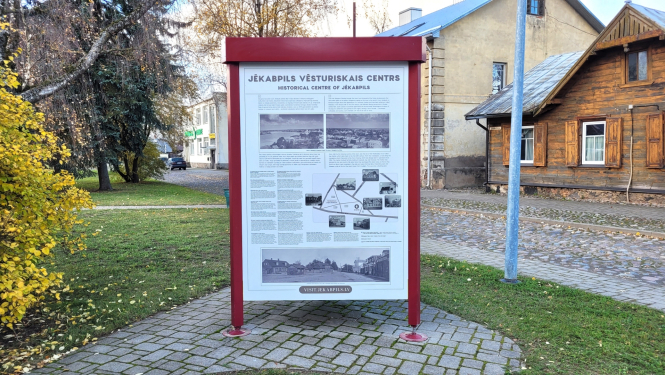 Gaišs trīs plakņu stends ar tumši sarkanu metāla konstrukciju uz bruģa