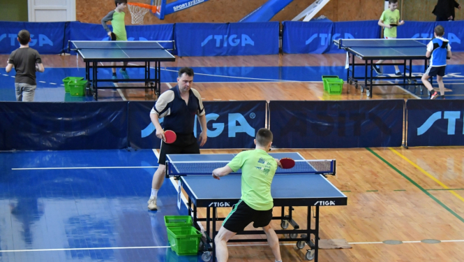 Divi vīrieši sporta zālē spēlē galda tenisu, fonā citi spēlētāji un galdi