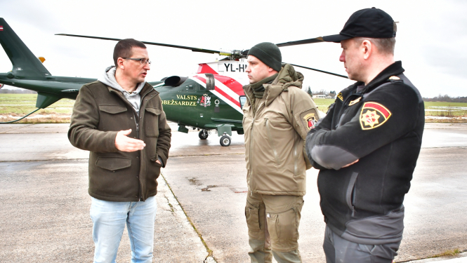 Domes priekšsēdētājs runā ar dienestu pārstāvjiem pirms lidojuma, fonā helikopters