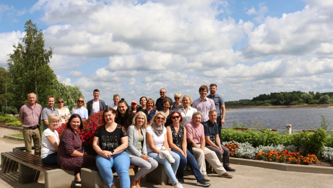 Pašvaldību dalībnieki grupā sēž uz soliņiem, fonā Daugava un puķu dobes