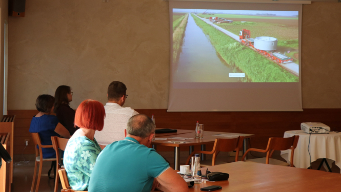 Cilvēki ar mugurām sēž pie galdiem un skatāvs video uz sienas