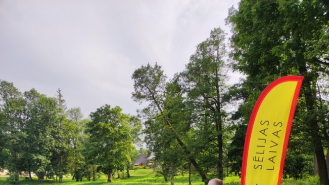 Laivotājs sporta tērpā stāv ūdens tilpnes krastā ar zāli, blakus zila kanoe laiva