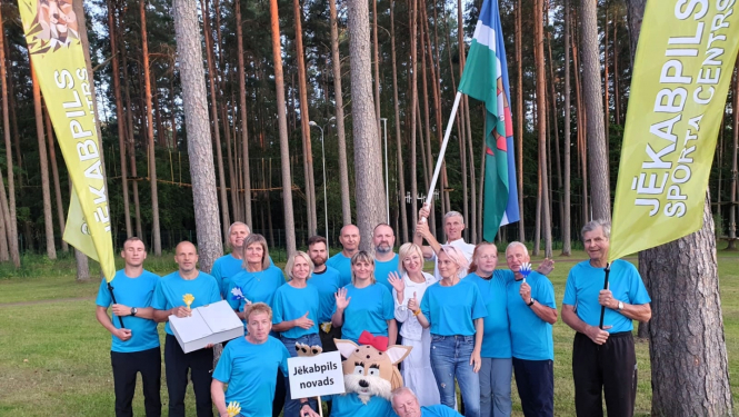 Jēkabpils novada vieglatlētu, kas piedalījās sacensībās, kopbilde ar ziliem krekliņiem, fonā novada karogs