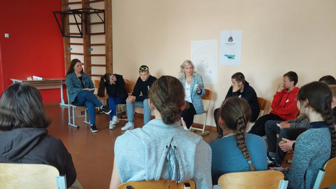 Projekta publicitātes foto - persona runā ar jauniešiem sēžot aplī krēslos