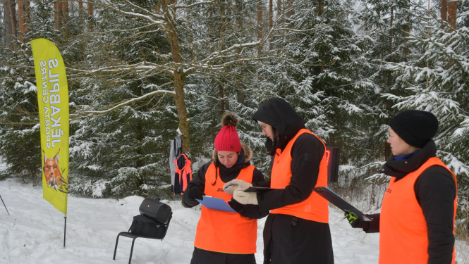 Pusstundas skrējiena ilustratīvs foto - cilvēki vestēs ziemīgā mešā skatās orientēšanās kartē