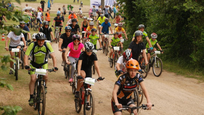 Liels daudzums velosipēdistu dažādos vecumos brauc pa zemes ceļu pretī fotogrāfam