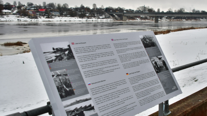 Ziemas skatā priekšplānā pelēks informatīvais stends ar tekstu dažādās valodās, fonā Daugava
