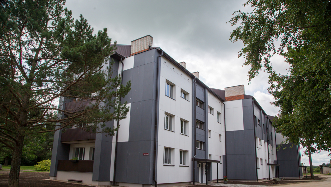 Trīs stāvu daudzdzīvokļu ēka ar jaunu pelēcīgu un baltu krāsu fasādi, lejā uzrušināta zeme, apkārt priedes