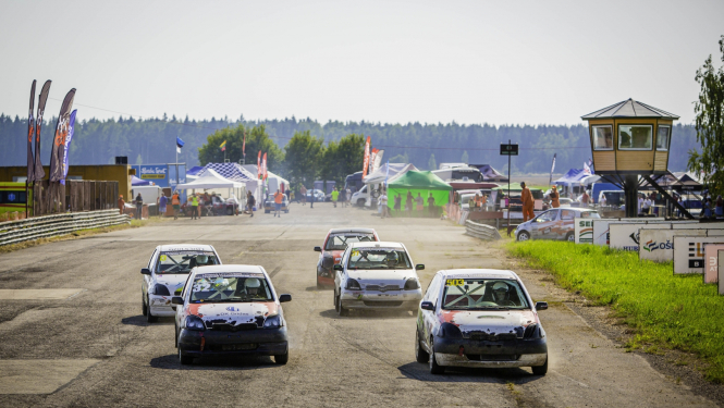 Rallijkrosa automašīnas dodas trasē, fonā tiesnešu būdiņa, teltis un skatītāji