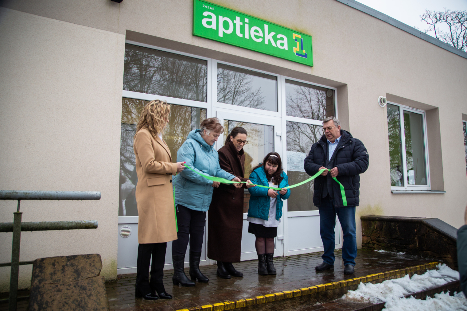 Svinīgā lentas griešana - stāv personas pie aptiekas ieejas durvīm