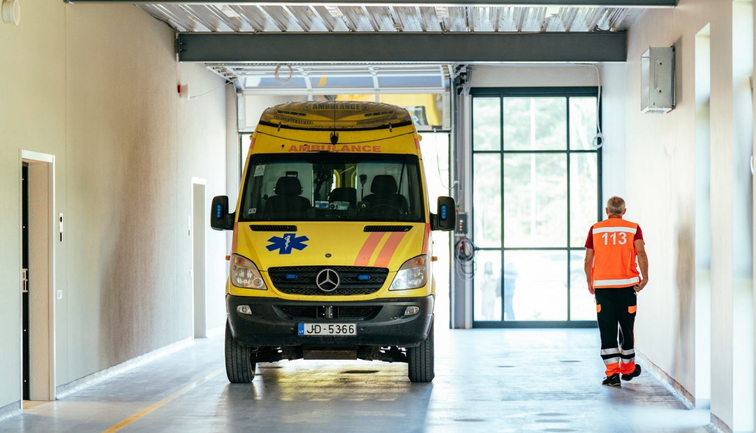 Dzeltena ātrās palīdzības mašīna telpās, blakus pa labi iet ātrās palīdzības darbinieks oranžā vestē