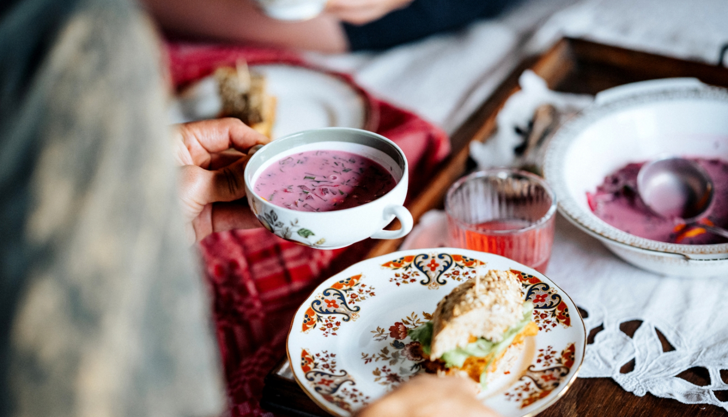 Persona pie galda no mugurpuses, sēdus, rokās tur šķīvi, fonā aukstās zupas bļodiņas
