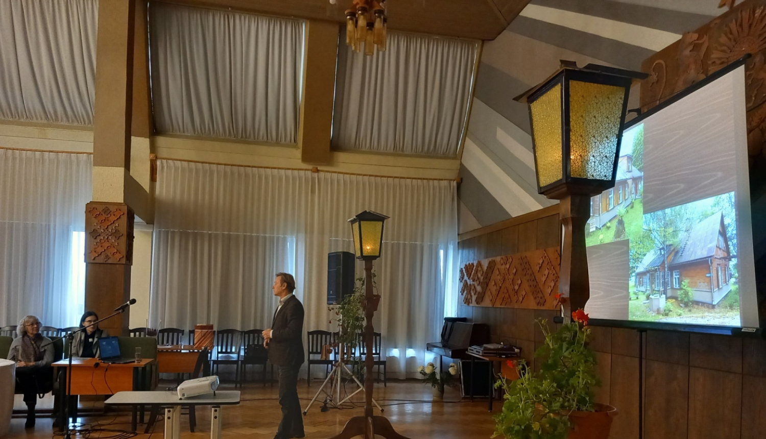 Persona runā lielā zālē, fonā projektorā rāda ēkas bildi