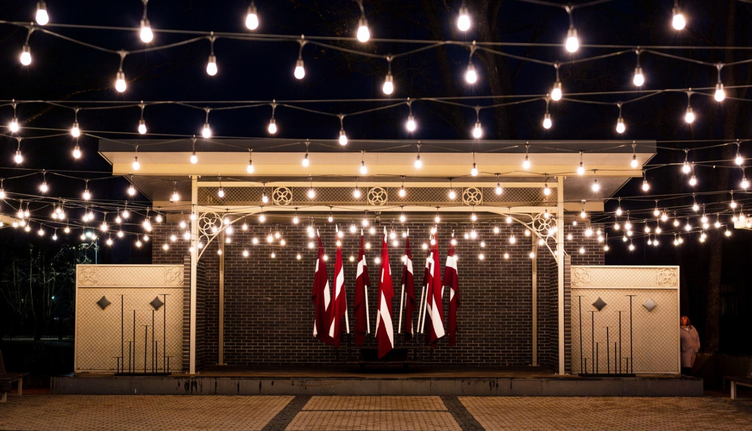 Kena parka estrāde naktī - izgaismota ar lampiņām un fonā Latvijas karogi