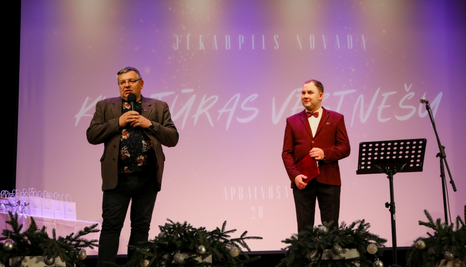 No kreisās Alfons Žuks ar vadītāju uz skatuves, fonā ekrāns ar uzrakstu Jēkabpils novada Kultūras vēstnešu apbalvošana
