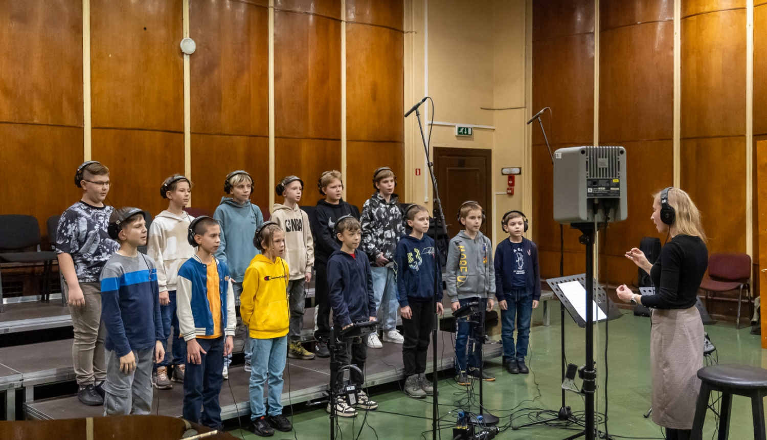 Zēnu koris sastājušies dzied un sieviete diriģē