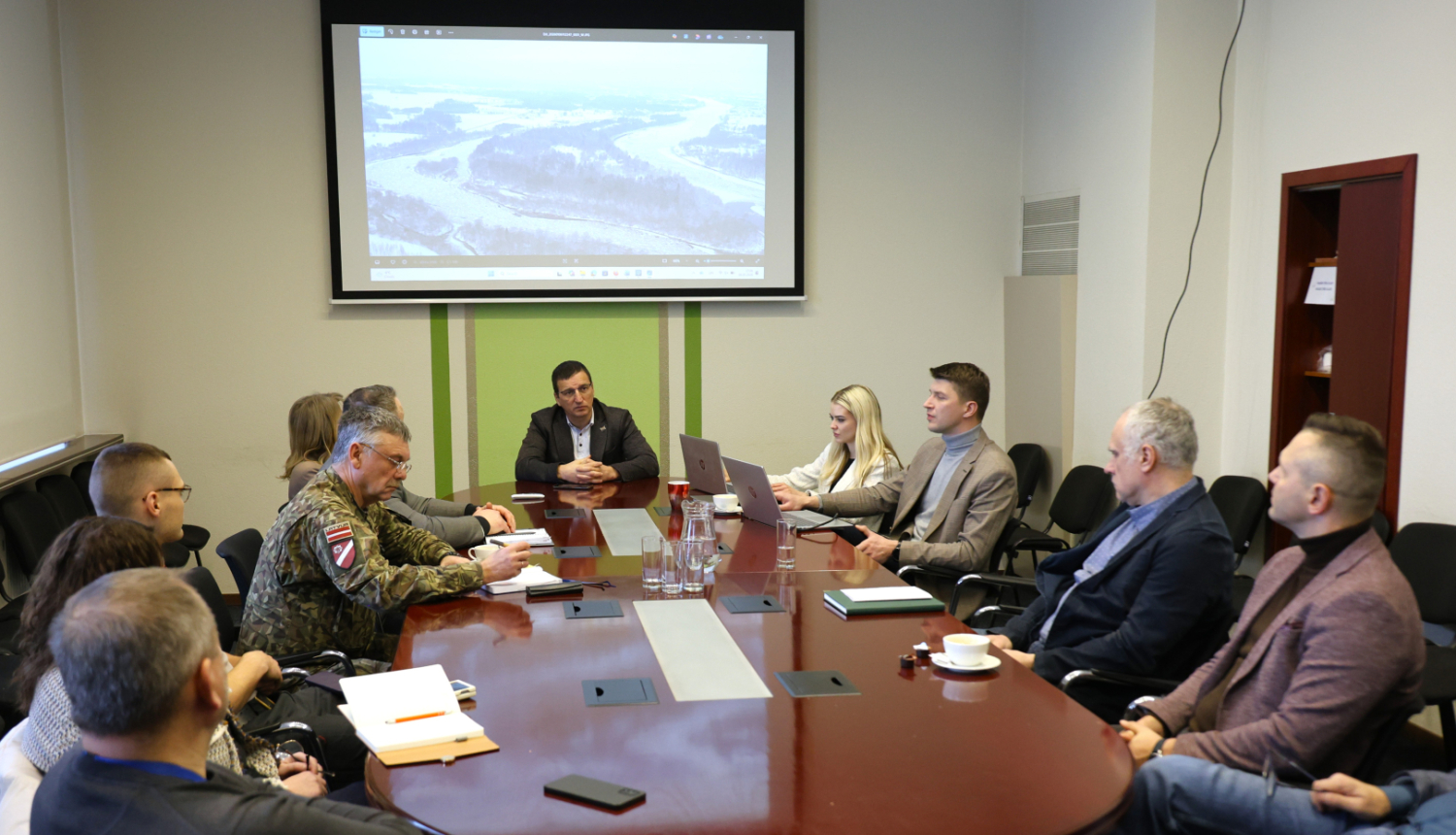 Komisija telpā pie galda, fonā ekrāns ar Daugavas skatu