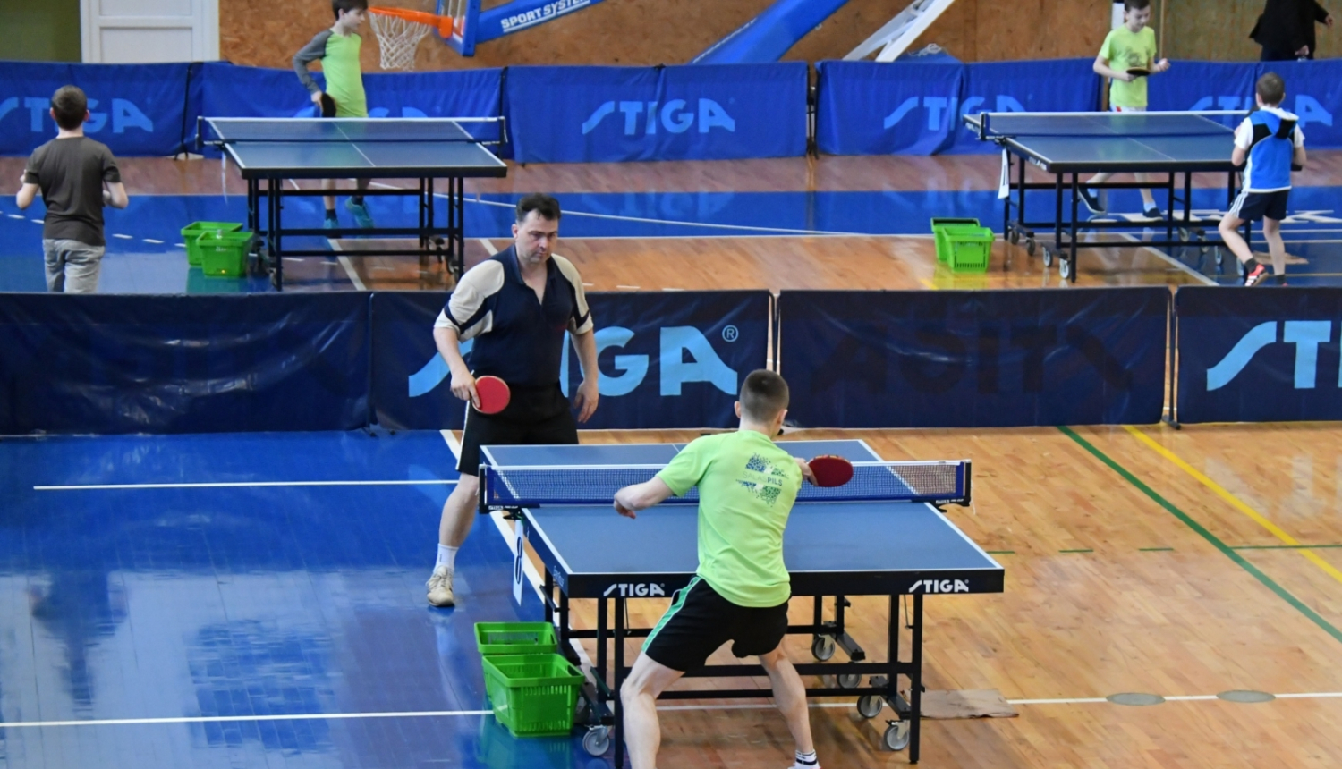 Divi vīrieši sporta zālē spēlē galda tenisu, fonā citi spēlētāji un galdi