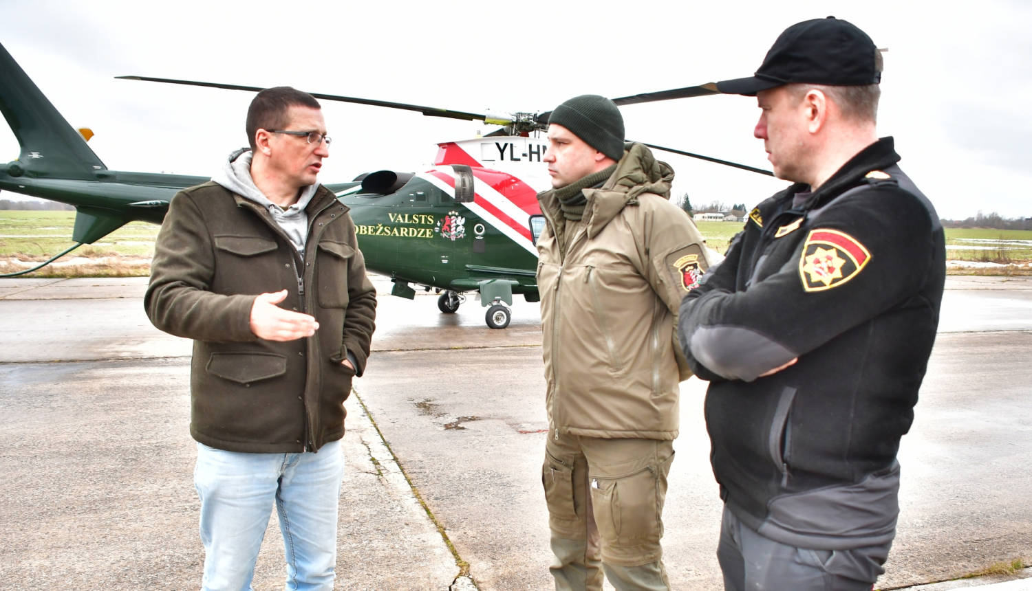 Domes priekšsēdētājs runā ar dienestu pārstāvjiem pirms lidojuma, fonā helikopters