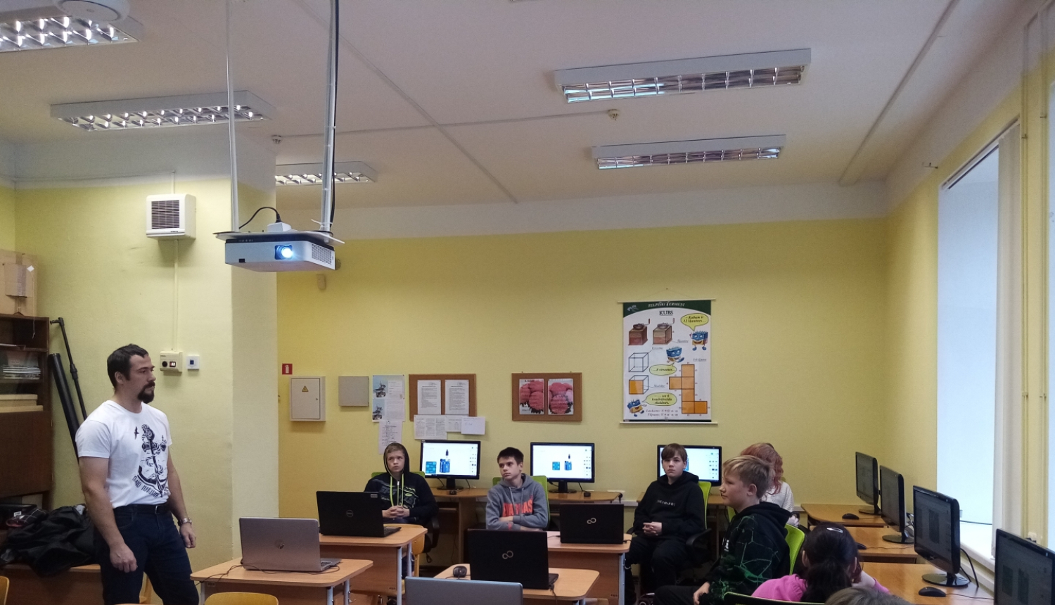 A.Bite (no kreisās) stāv un māca skolēnus, kas sēž klasē pie datoriem