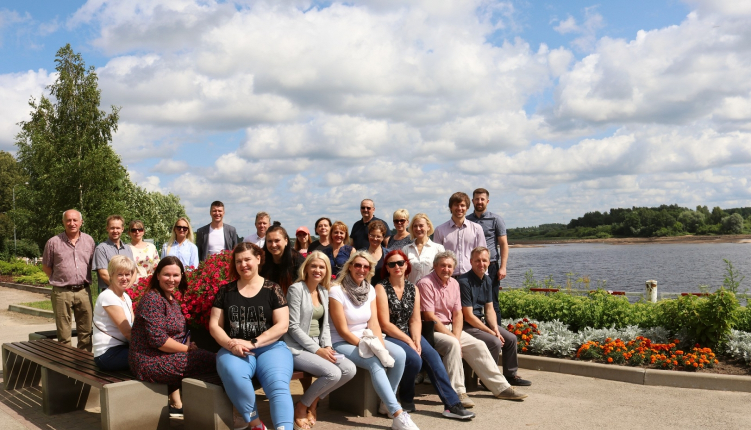 Pašvaldību dalībnieki grupā sēž uz soliņiem, fonā Daugava un puķu dobes
